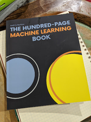 Photograph of the paperback copy of The Hundred-Page Machine Learning Book by Andriy Burkov lying on a grid-paper notebook on a wooden desk