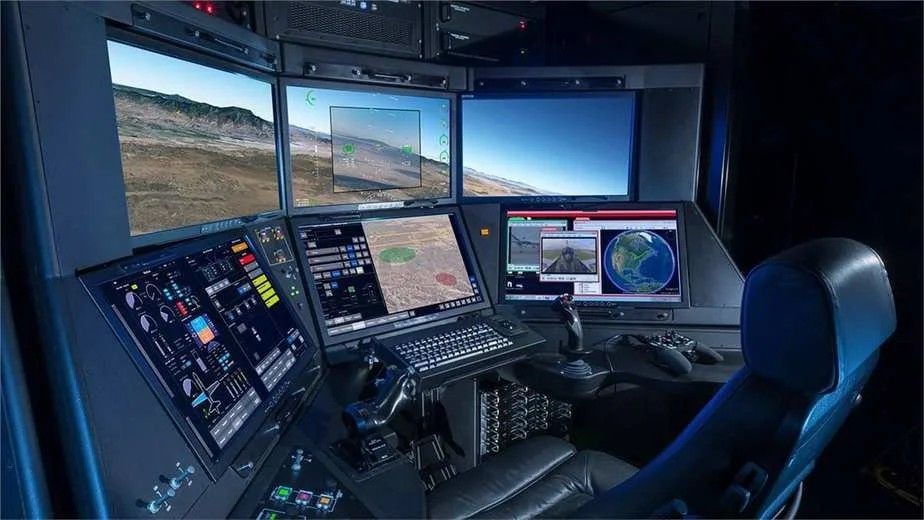 Darkened unmanned-aircraft ground control station: curved array of six displays around a black leather operator seat showing flight view over arid terrain, aircraft status, tactical map, and runway view, with twin joysticks and throttle